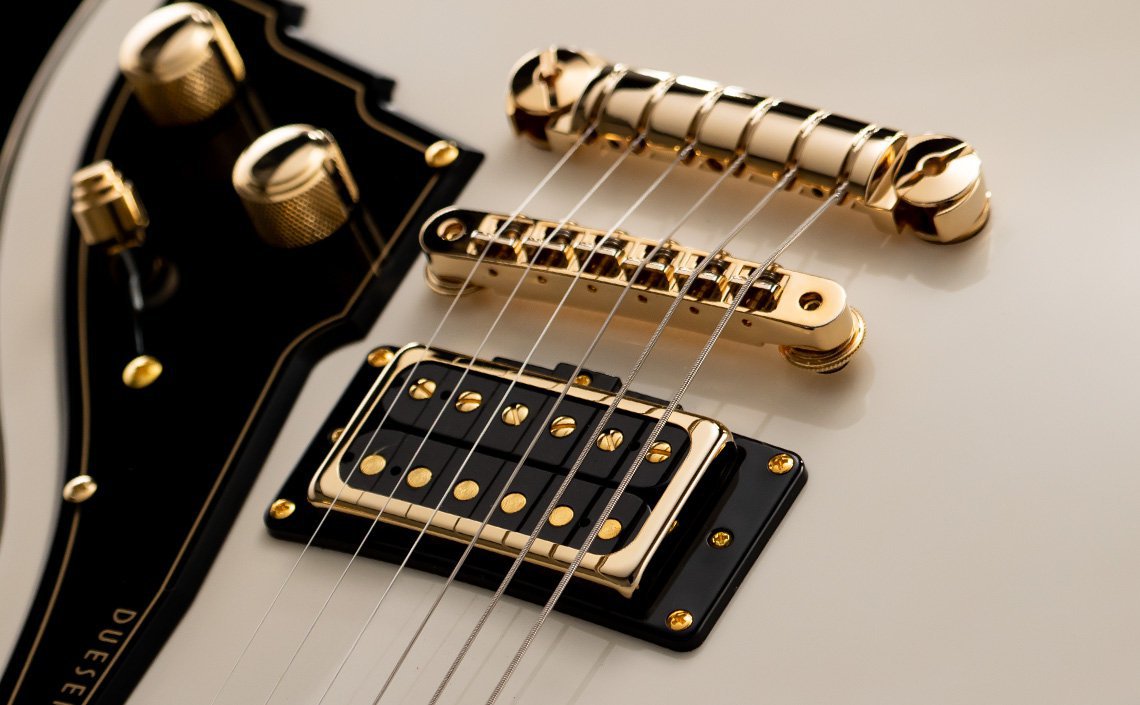 Close-up of a Duesenberg Fantom A electric guitar in a sleek white finish with gold hardware. The image highlights the black pickguard with gold screws, humbucker pickups, metallic strings, and control knobs. The luxurious design emphasizes precision craftsmanship and premium style.