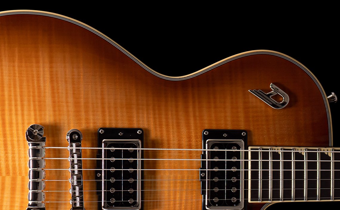 A close-up of an electric guitar with a vibrant sunburst finish, blending shades of orange and brown. The focus is on the bridge, pickups, strings, and part of the fretboard, showcasing the instrument's meticulous craftsmanship and stylish design. The iconic Duesenberg logo is also visible on the body, adding a touch of elegance and identity.