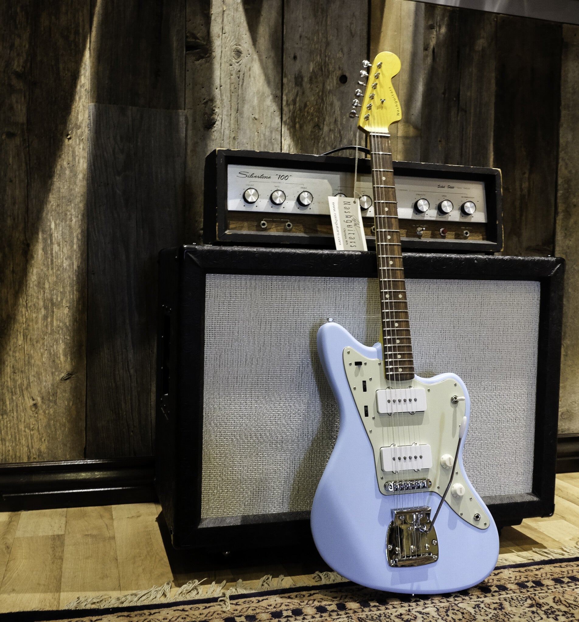 Front of a light blue Nash electric guitar with a pickguard, leaning against a vintage Silvertone '100' amplifier.