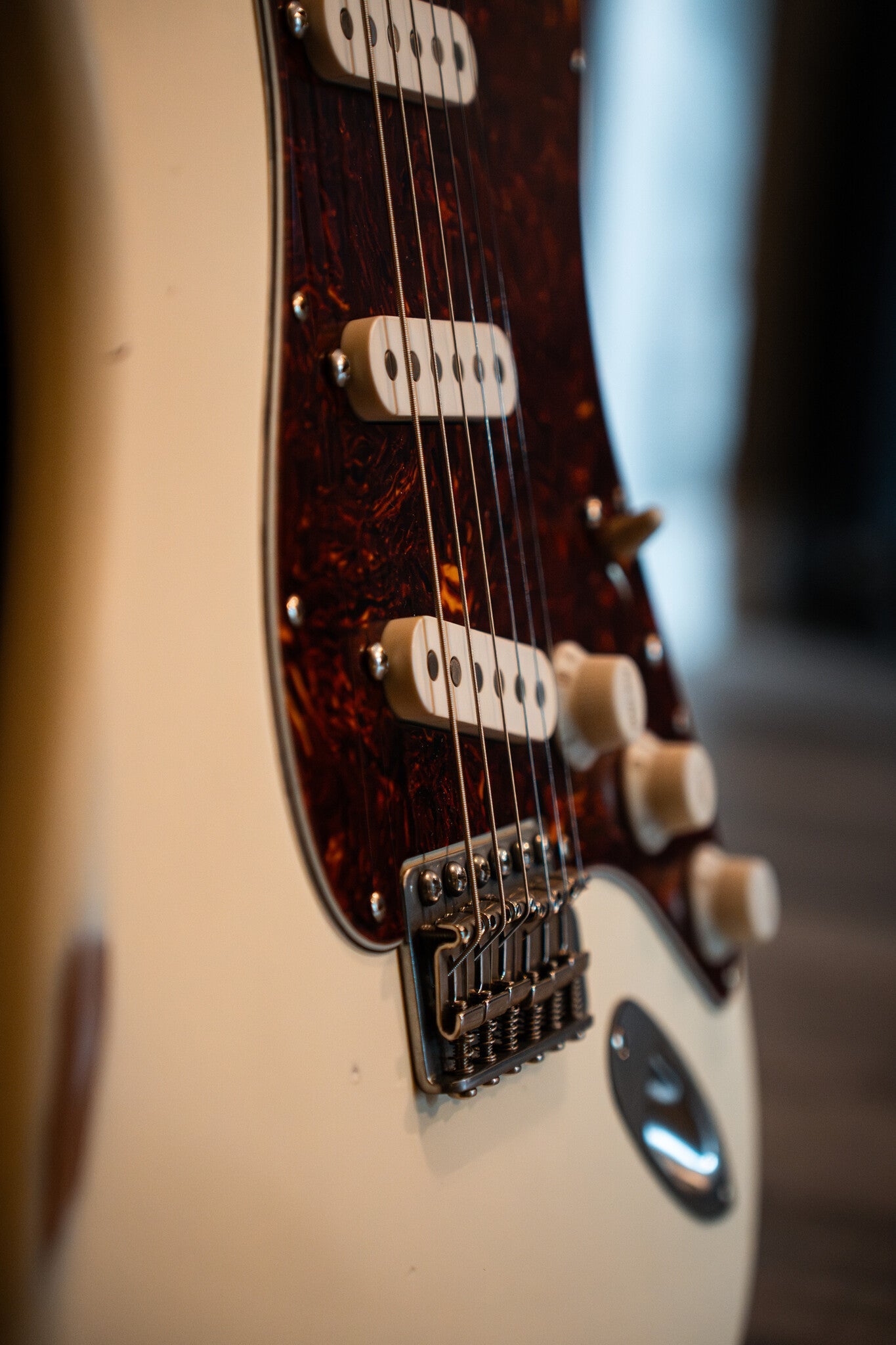 Close-up of a cream-colored electric guitar with a tortoiseshell pickguard, three single-coil pickups, metallic bridge components, and control knobs.