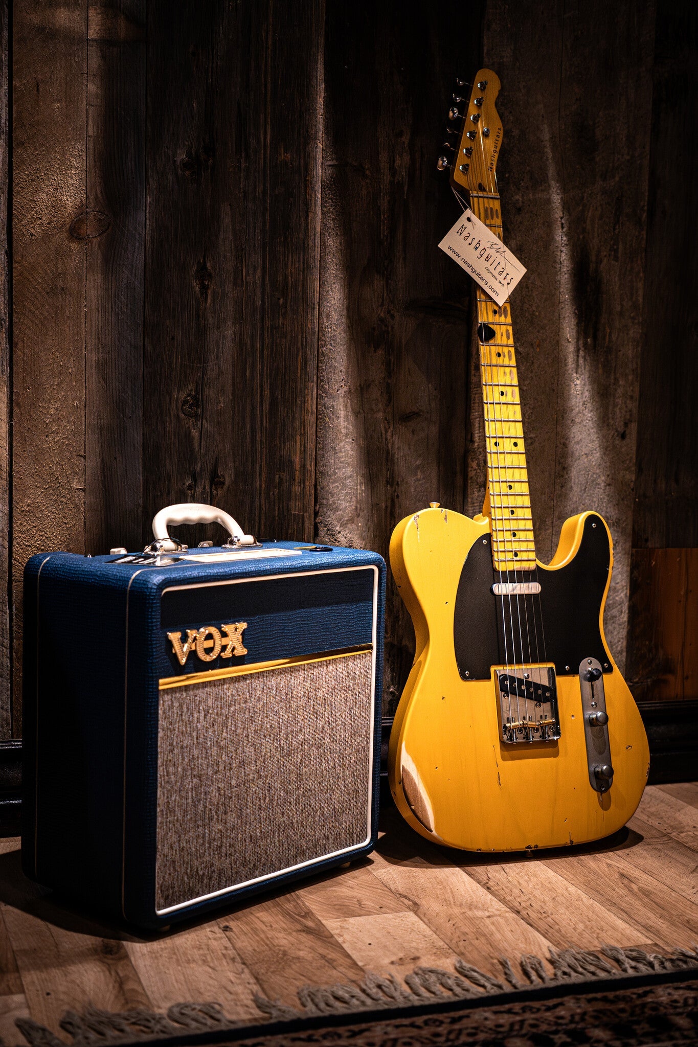 Yellow Nash electric guitar leaning against a curtain wall, next to a blue and gray VOX amplifier.