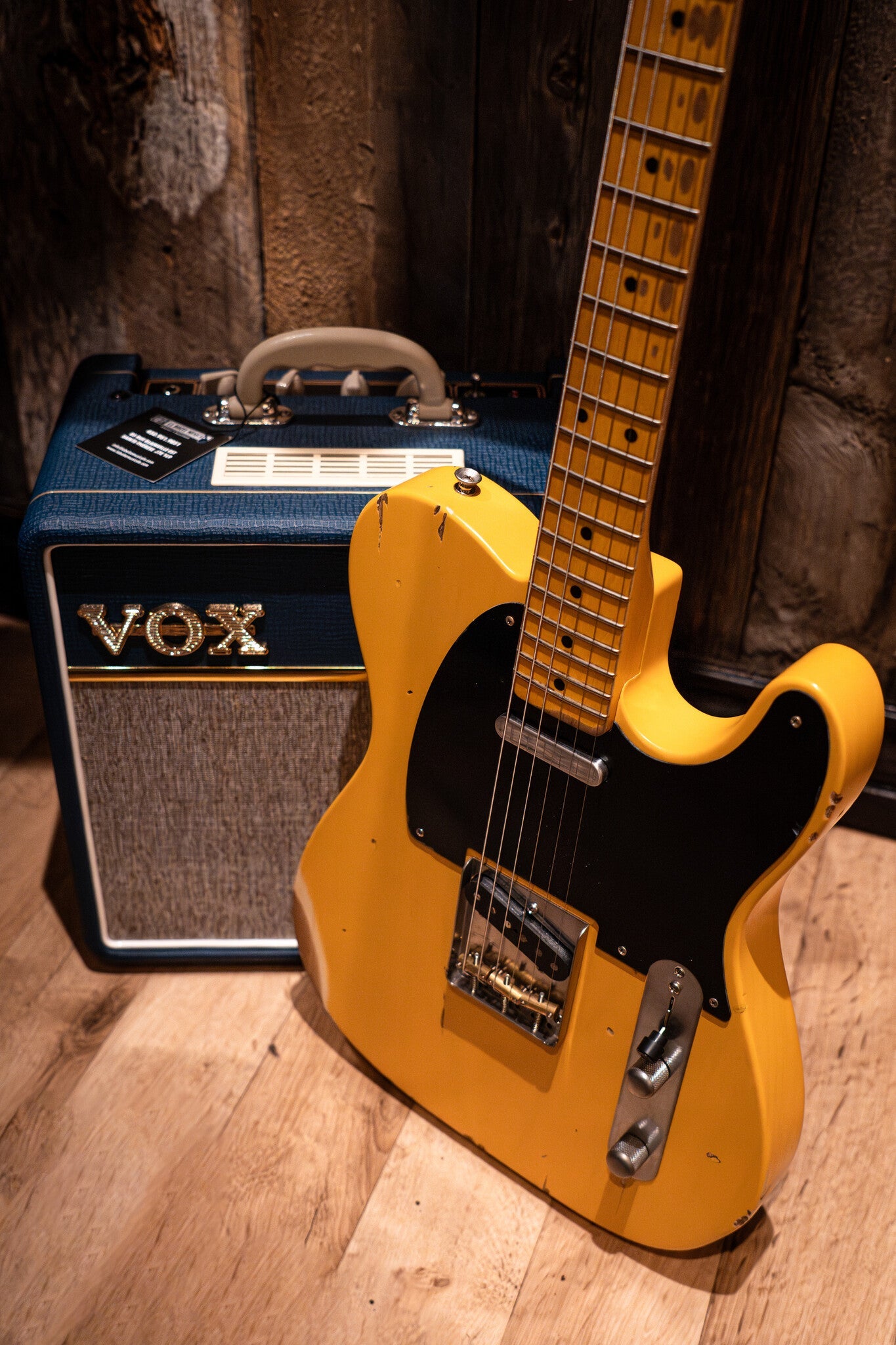 Yellow Nash electric guitar with a black pickguard, featuring visible wear and relic-style aging, leaning against a blue and gray VOX amplifier with a white handle.
