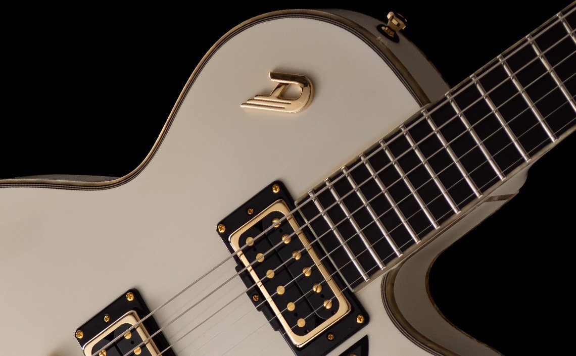 Close-up of a Duesenberg electric guitar in a white finish with elegant gold hardware. The focus is on the intricate details, including black and gold pickups, metallic strings, and the iconic gold 'D' logo on the body. The fretboard with metal frets and a dark finish further highlights the guitar's sophisticated design and premium craftsmanship.